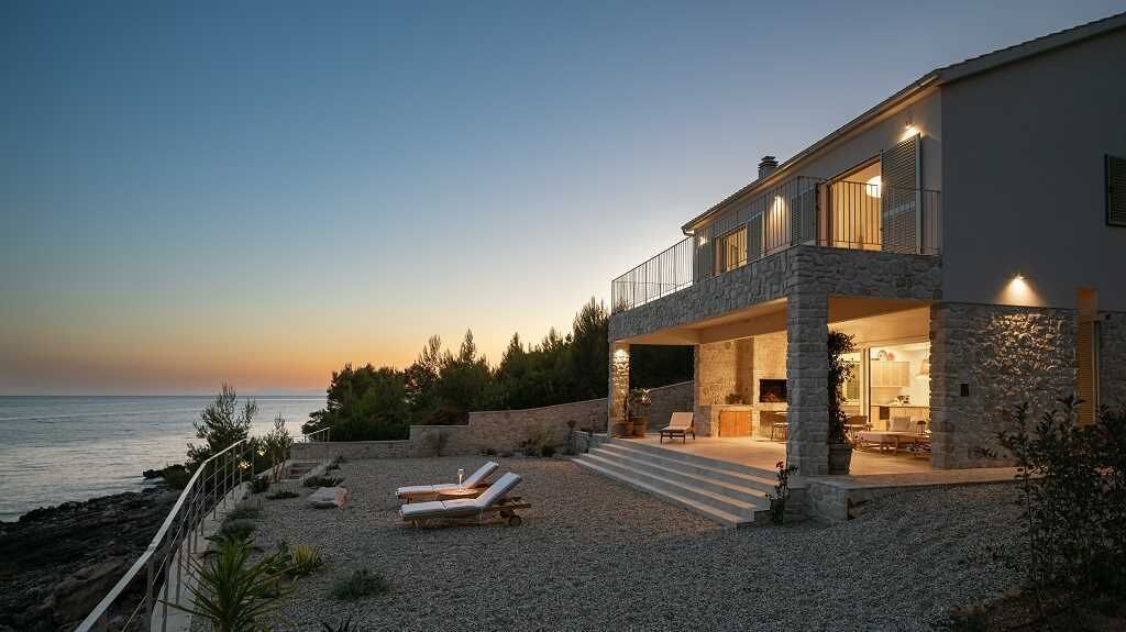 A coastal house with a stone facade, a balcony, and a pool area with lounge chairs.