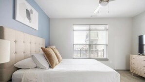 Un dormitorio con cama, ventilador de techo, ventana con persianas y televisor sobre un tocador.