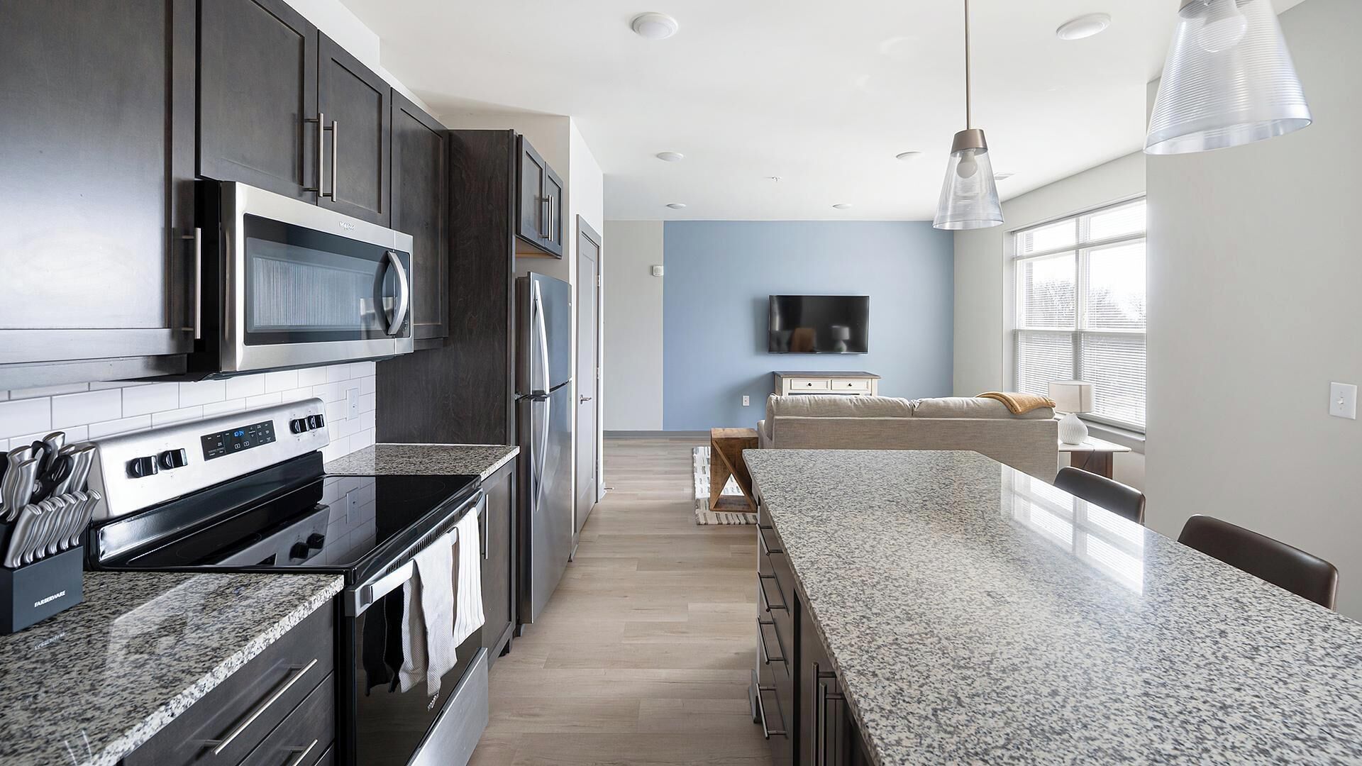 A modern kitchen with granite countertops, stainless steel appliances, and a flat-screen TV mounted on the wall.