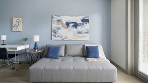 Una sala de estar moderna con un sofá gris, dos almohadas azules, una mesita de noche blanca, una lámpara azul y un cuadro abstracto enmarcado en la pared.