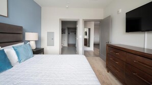Un dormitorio con cama, mesita de noche, lámpara, cómoda, televisor y un baño visible a través de una puerta abierta.