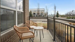 A balcony with wicker chairs and a small table, overlooking a street with buildings and a clear sky. - Landing | Modern 2BD, Gym, Pool (Nashville)