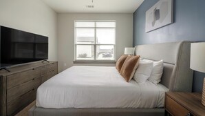 A bedroom with a bed, a TV on a wooden dresser, a window with blinds, and a wall-mounted lamp. - Landing | Modern 2BD, Gym, Pool (Nashville)
