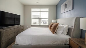 A bedroom with a bed, a TV on a wooden dresser, a window with blinds, and a wall-mounted lamp. - Landing | Stylish 2BD, Clubhouse, Pool (Nashville)