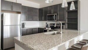 A modern kitchen with dark cabinets, stainless steel appliances, granite countertops, and pendant lighting. - Landing | Modern 2BD, Gym, Pool (Nashville)