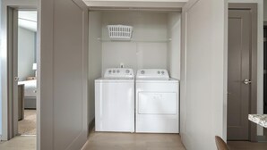 A laundry room with two white front-loading washers and dryers, a white laundry basket on the shelf, and a door leading to another room. - Landing | Modern 2BD, Gym, Pool (Nashville)