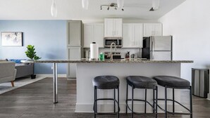 A modern kitchen with white cabinets, stainless steel appliances, and a granite countertop. There are three bar stools and a dining area with a grey sofa.