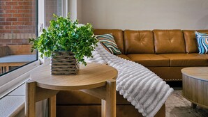 A living room with a brown leather sofa, a white textured throw blanket, a round wooden table with a potted plant, and a framed artwork on the wall. - Sleeps 12 · Game Room · Foosball · Yard BBQ · 3BR Near Anschutz & Denver (Aurora)