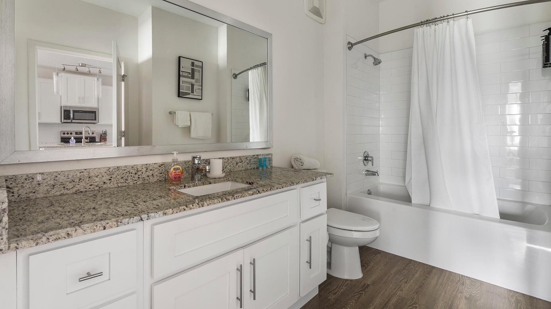 Un baño con encimera de granito, muebles de color blanco, un inodoro, ducha con cortina y un espejo grande.
