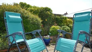 Two turquoise lawn chairs with metal frames, positioned on a patio with a garden and a blue pot in the background. - Bumble Cottage - Parking - Pet - Garden - Sleeps 4 (Ilfracombe)