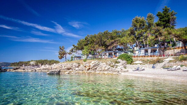 A coastal hotel with clear waters, rocky shoreline, and pine trees.