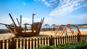 A wooden ship-shaped play structure on a sandy beach with a wooden fence in the foreground.