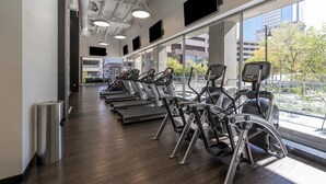 Un gimnasio con cintas de correr y máquinas elípticas, ventanas amplias y vistas a edificios y árboles.