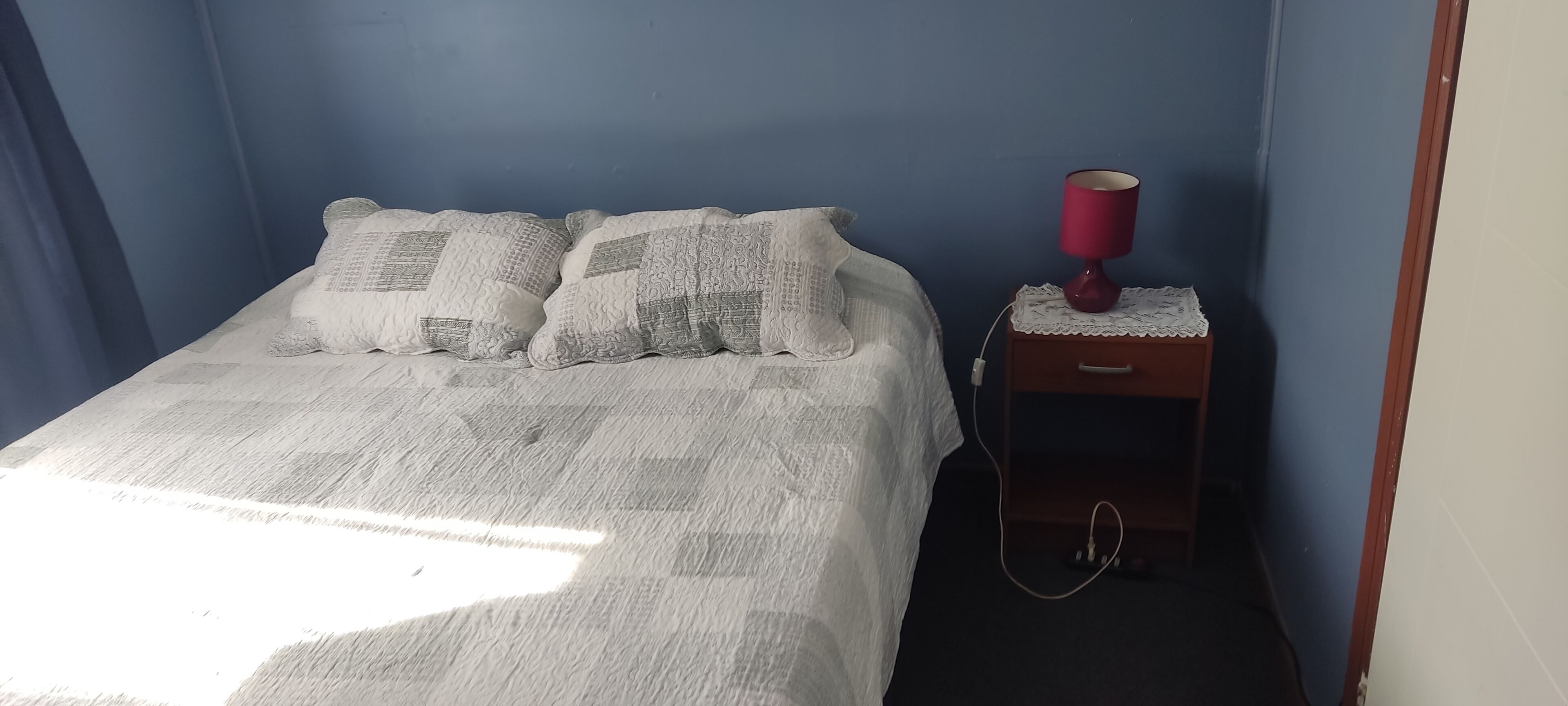 A bedroom with a bed, pillows, a nightstand with a lamp, and a power outlet.