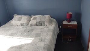Een slaapkamer met een bed, kussens, een nachtkastje met een lamp en een stopcontact.
