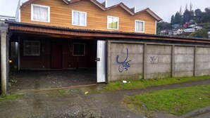 A two-story wooden house with a garage, a concrete wall with graffiti, and a grassy area. - C1 Furnished Cabin Near downtown Puerto Montt (Puerto Montt)