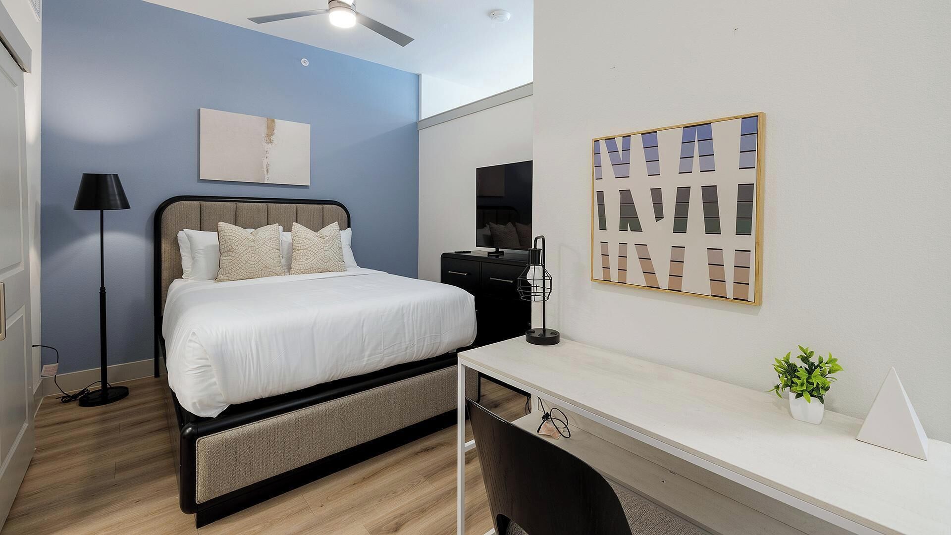 Een slaapkamer met een bed, nachtkastje, bureau, stoel, lamp, kunstwerk en een plafondventilator.