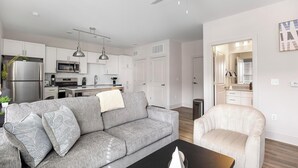 A modern living room with a grey sofa, a white armchair, a black coffee table, and a kitchen in the background. - Landing | Modern 2BD, Game Room, Courtyard (Richmond)