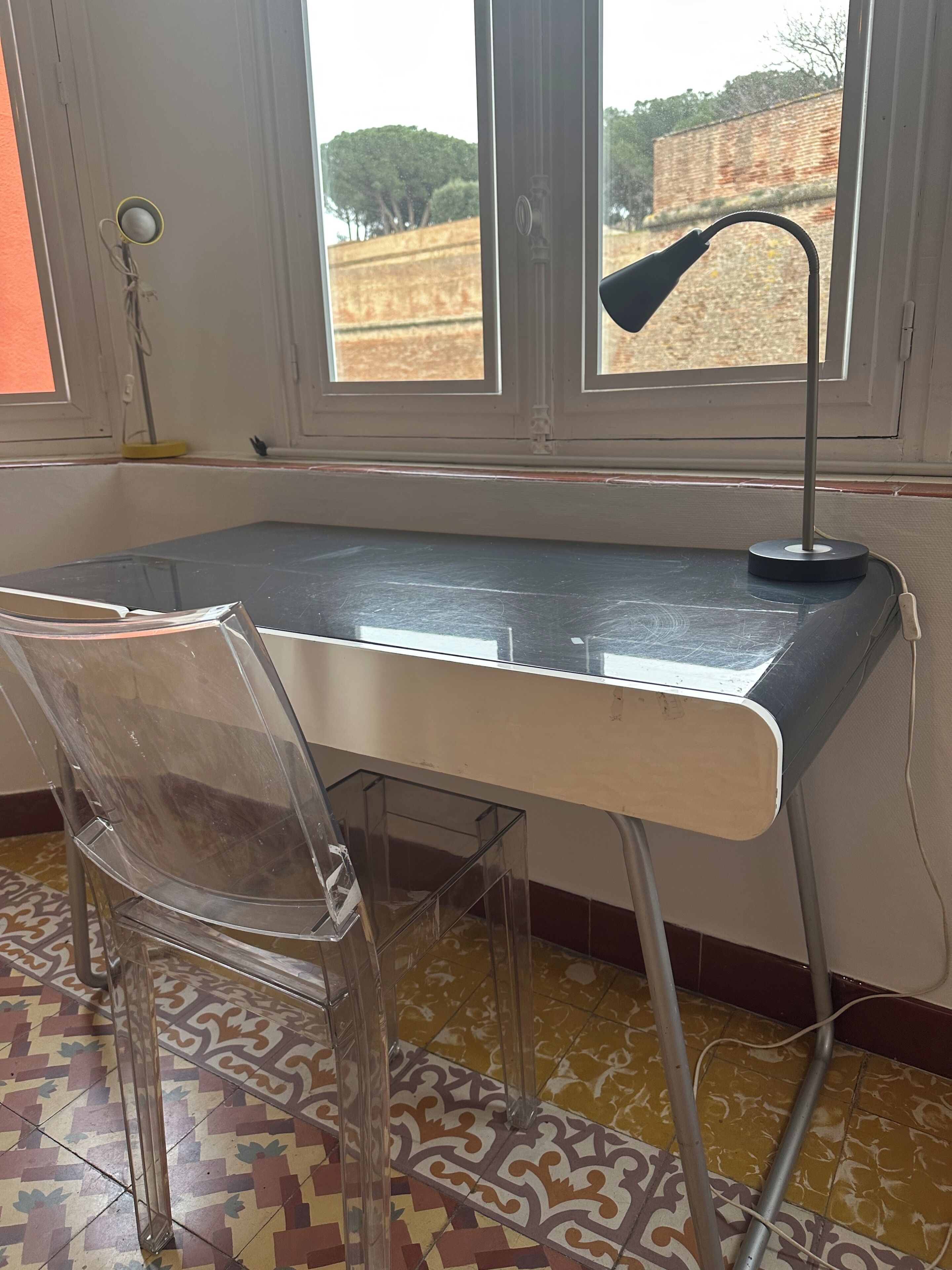 A room with a desk, a chair, a window with a view of trees and a brick wall, and patterned floor tiles.