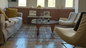 A room with a sofa, armchair, and a small table with decorative items. There are three windows with a view of greenery outside. - Apartment Perpignan center 92 m2, 2 bedrooms 2 bathrooms, parking (Perpignan)