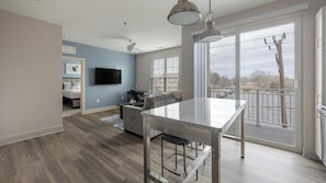 A modern living room with a flat-screen TV, a sofa, a dining table, and a balcony with a view. - Landing | Incredible 1BD, Game Room, Gym (Richmond)