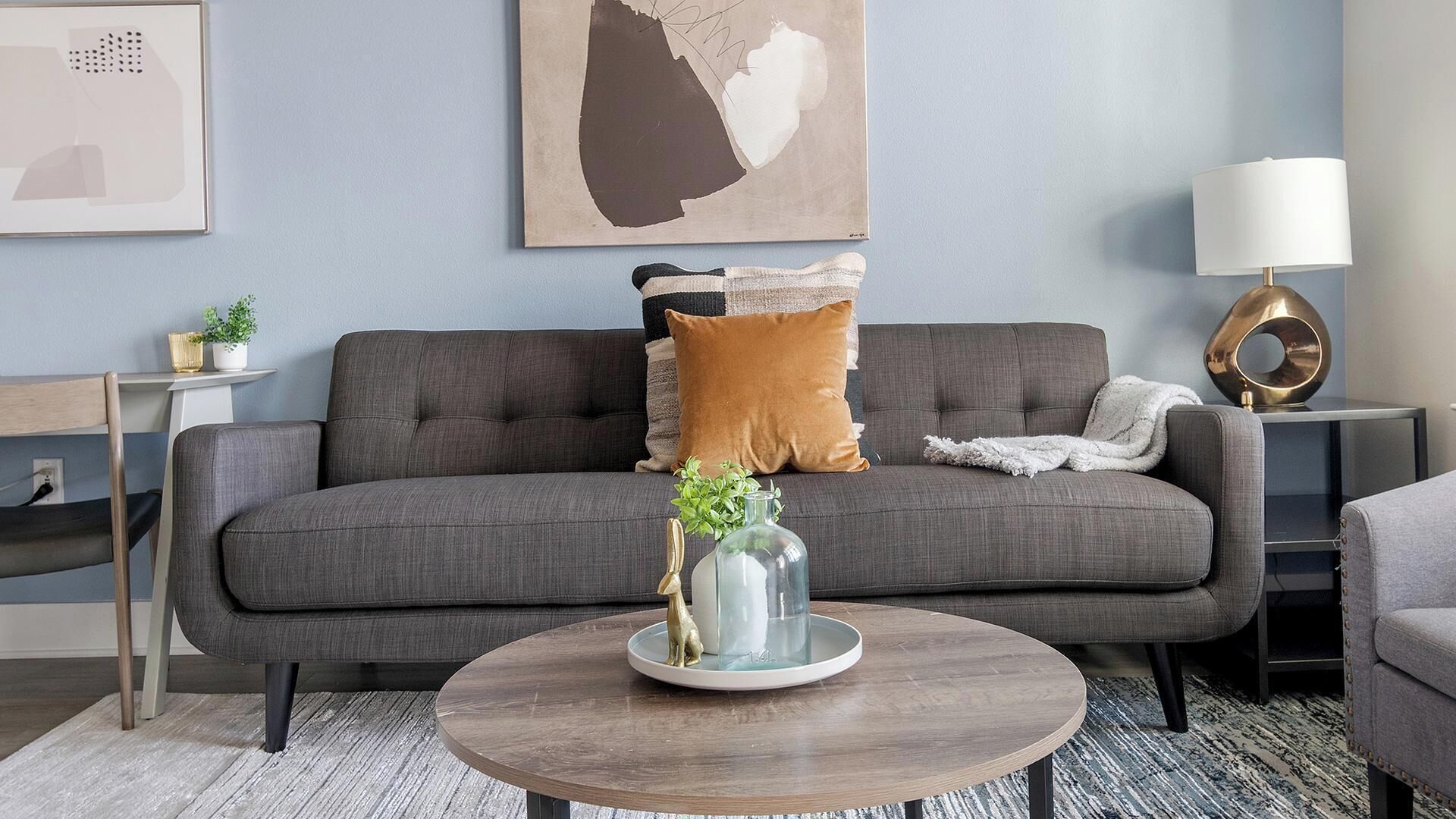 Un salon avec un canapé gris, une table basse ronde, une lampe et une œuvre d’art murale.