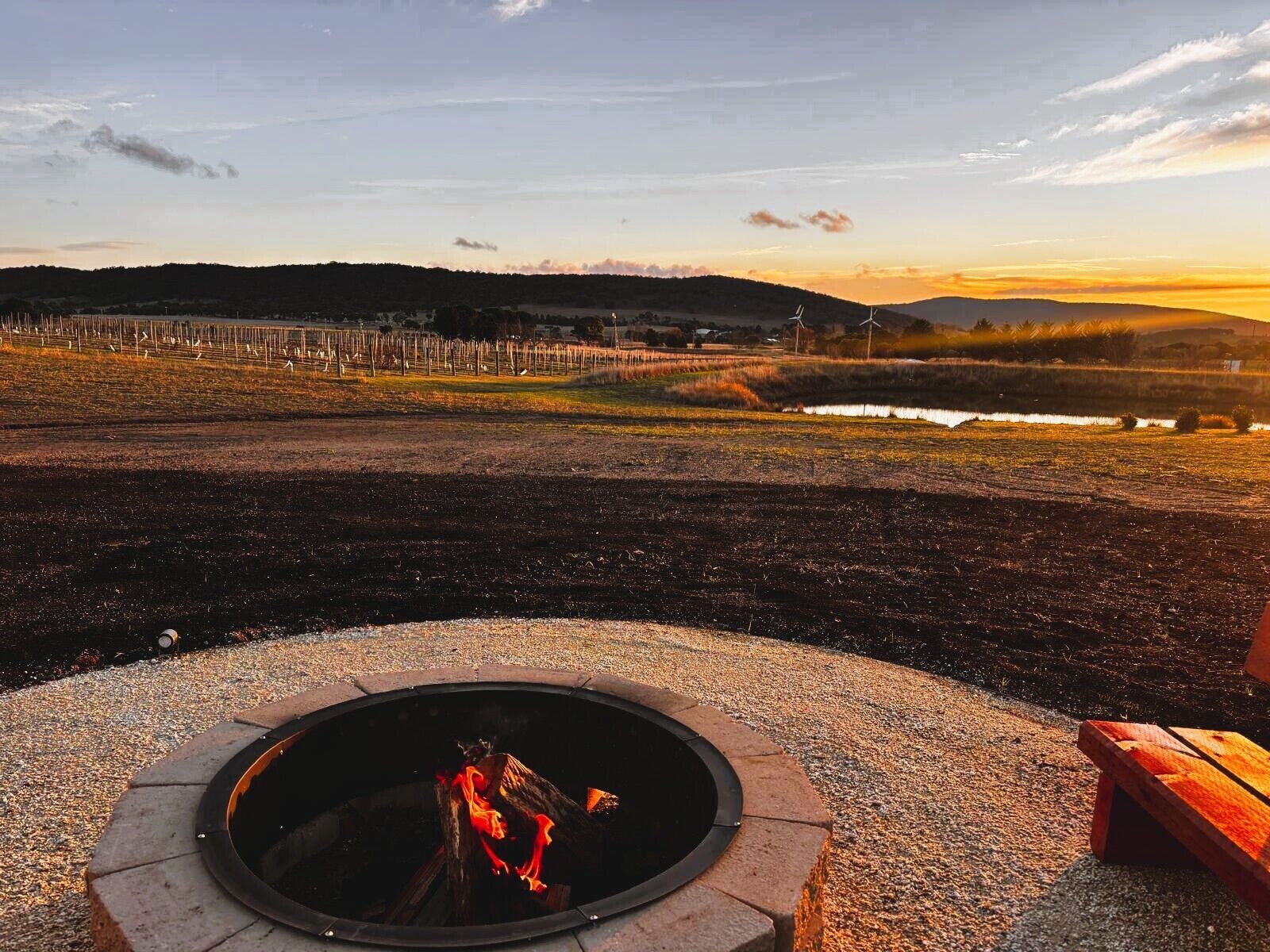 A fire pit with logs, surrounded by a gravel area, overlooking a vineyard and distant hills.