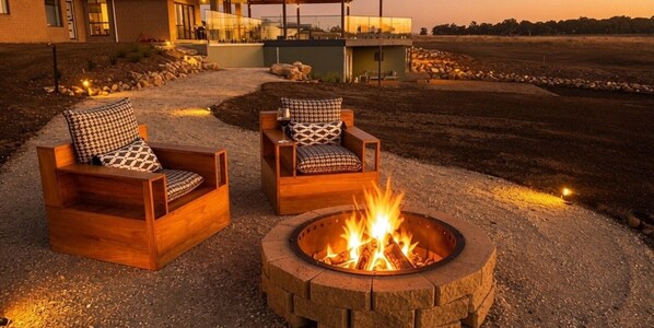 Two wooden chairs with patterned cushions, a circular fire pit with flames, and a building in the background.
