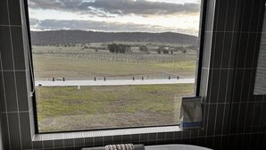 Une salle de bain moderne avec une baignoire sur pieds, une fenêtre donnant sur un champ et des collines, et des murs recouverts de carreaux foncés.
