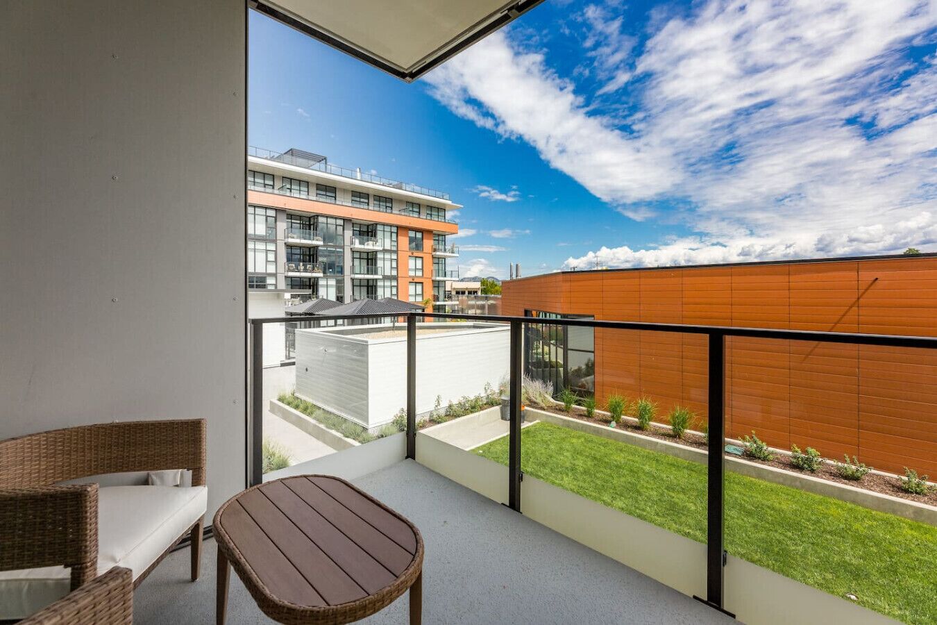 Un balcón con una silla de mimbre y una mesita pequeña, con vista a un edificio moderno y una zona verde.