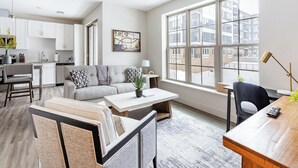 Ein modernes Wohnzimmer mit einer Couch, einem Couchtisch und Sesseln, eine Küche mit weißen Schränken und große Fenster mit Blick auf Gebäude.