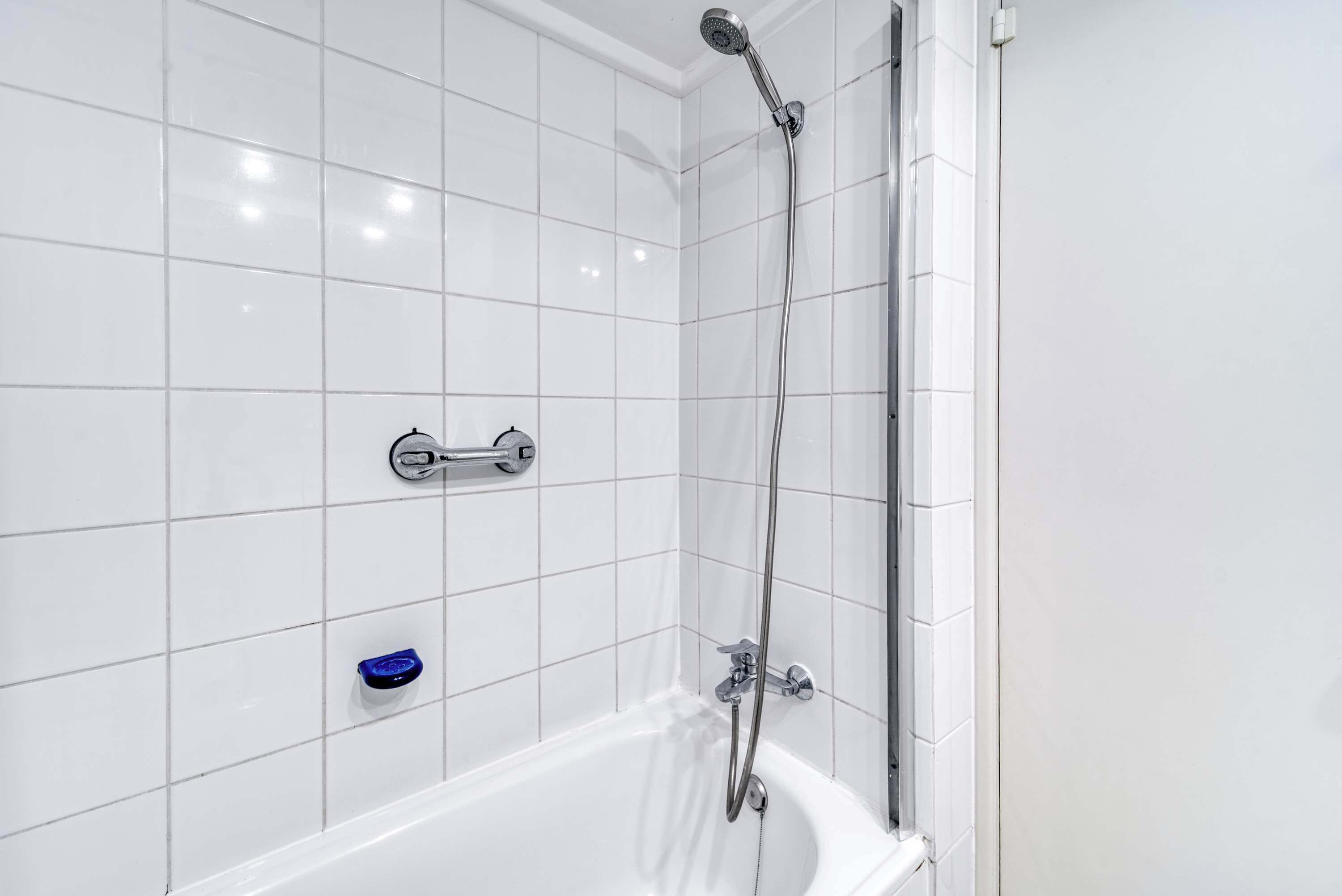 Ein Badezimmer mit weißen Fliesen, einer weißen Badewanne, einer Handbrause und einer kleinen, blauen Seifenschale.