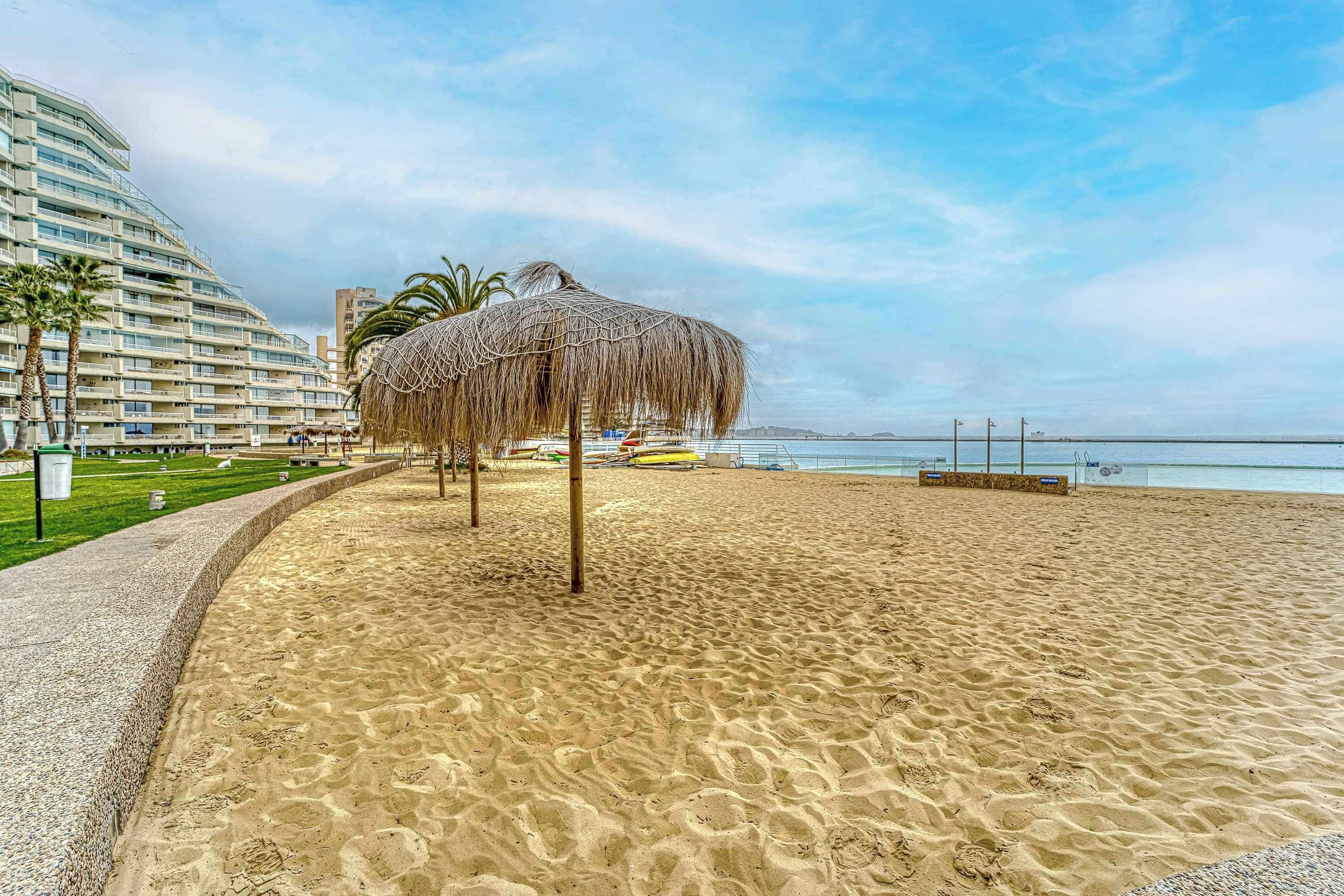 Une plage avec un parasol en chaume, un hôtel en arrière-plan et un ciel dégagé.