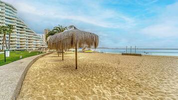 Une plage avec un parasol en chaume, un hôtel en arrière-plan et un ciel dégagé.