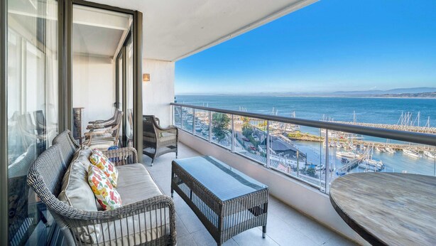 Un balcón con mobiliario de mimbre, una mesa de cristal y vistas al mar y al puerto deportivo.
