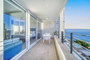 Un balcón con vista al mar, una zona de estar con una mesa y sillas, y un ventanal que da a una habitación con una cama y un sofá azul.