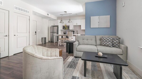 Una sala de estar moderna con un sofá, un sillón y una mesa de centro, una cocina al fondo y una pared azul de acento.