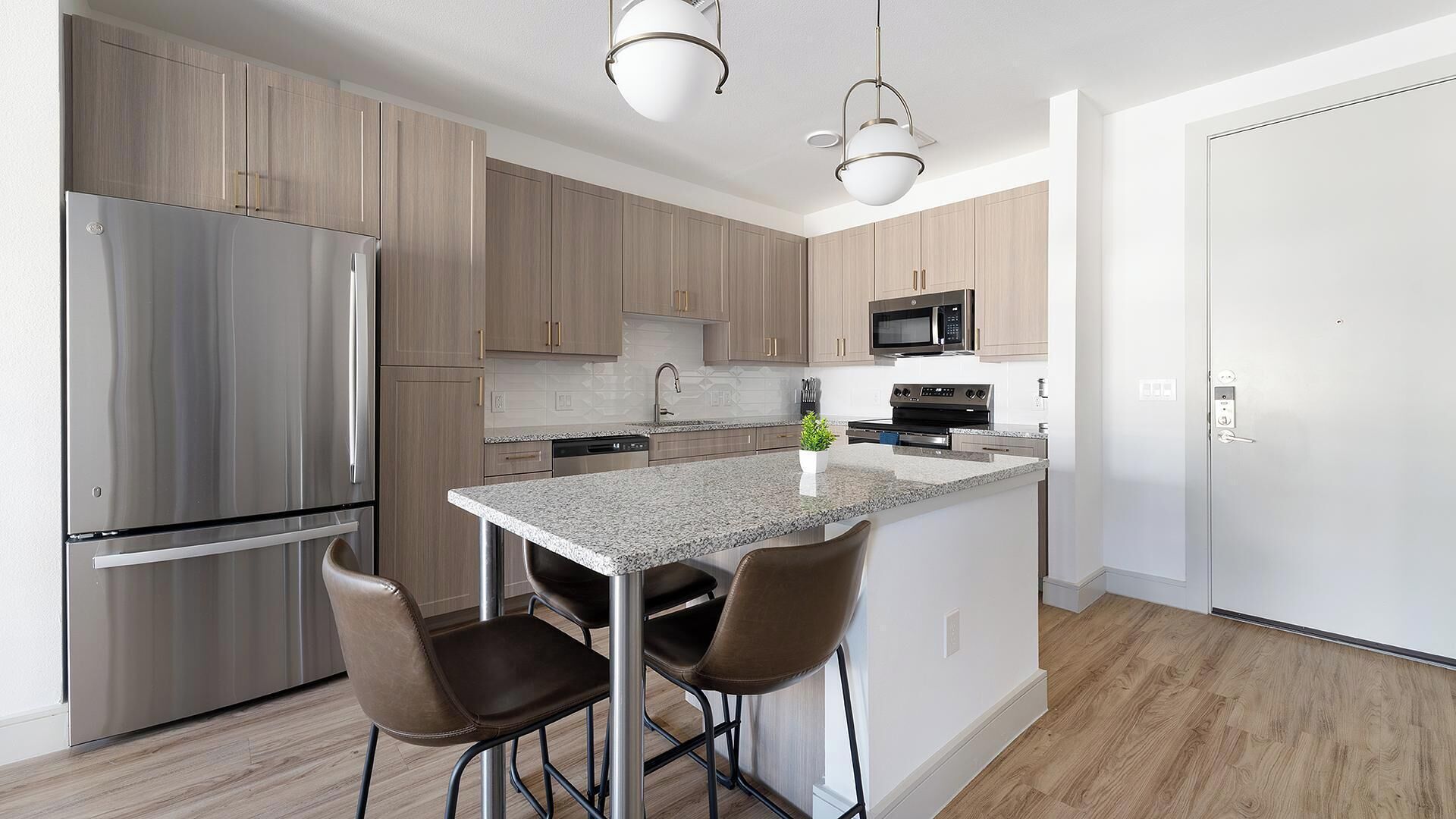 A modern kitchen with stainless steel appliances, wooden cabinets, a granite countertop, and two bar stools.