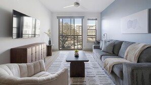 A modern living room with a sofa, a coffee table, a TV, and a balcony with a view of buildings. - Landing | Spectacular 1BD, Clubhouse, Pool (Austin)