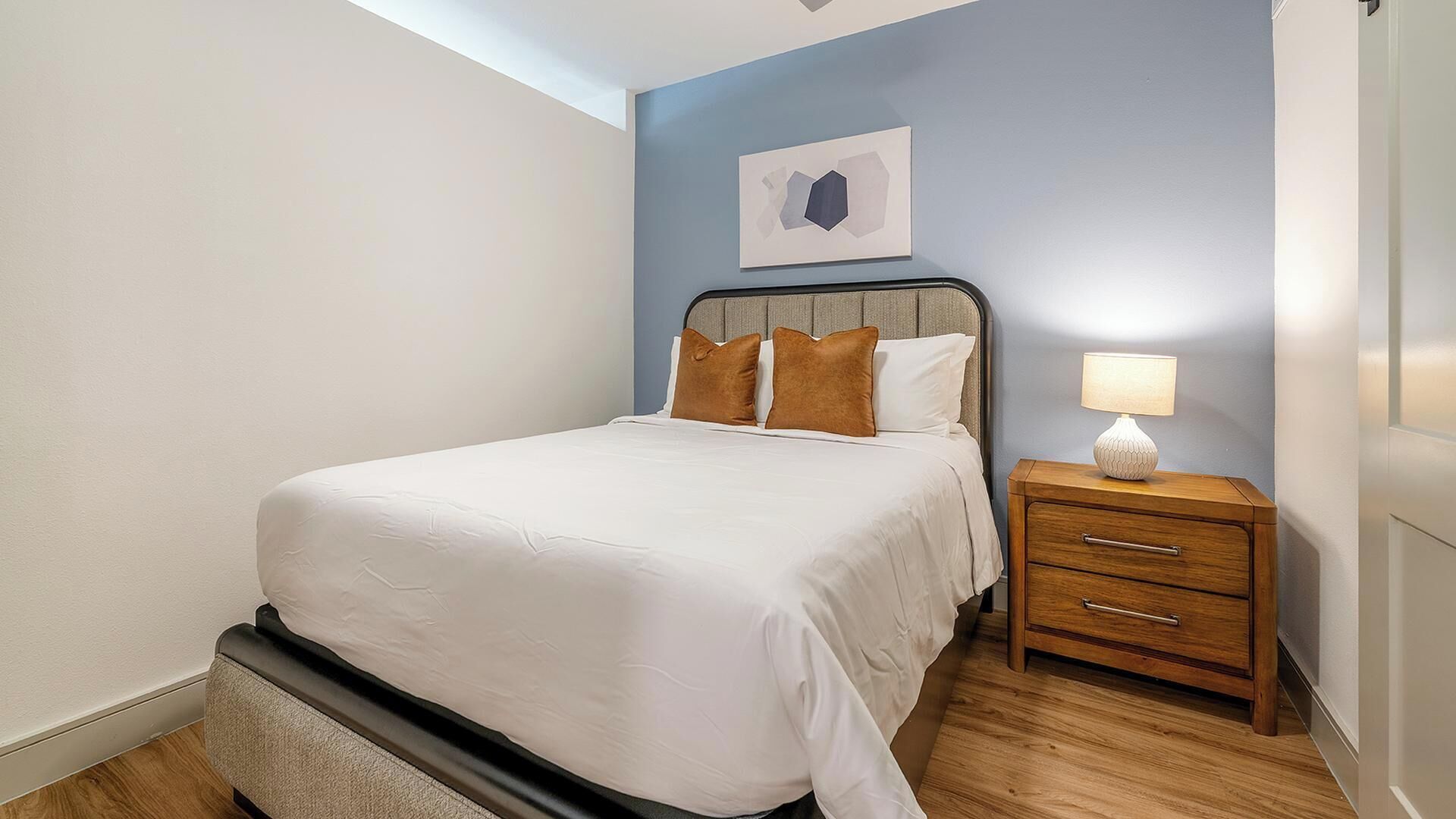 Una cama bien hecha con cabecera, una mesita de noche con lámpara y una obra de arte colgada en la pared.
