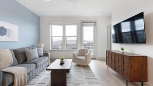 A modern living room with a sofa, armchair, coffee table, TV, and a view through large windows. - Landing | Beautiful 1BD, Pool, Clubhouse (Austin)