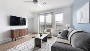 A modern living room with a flat-screen TV, a wooden cabinet, a grey sofa, a coffee table, and a large window with a view. - Landing | Beautiful 1BD, Pool, Clubhouse (Austin)