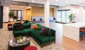 Ein modernes Wohnzimmer mit einem grünen Ecksofa, einem runden Couchtisch und einer Küchenzeile mit weißer Arbeitsplatte und hängenden Pendelleuchten.