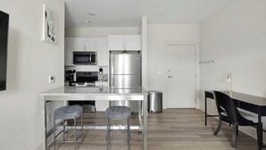 A modern kitchen with white cabinets, a stainless steel refrigerator, a microwave, and a coffee maker. There are two bar stools and a dining table with chairs. - Landing | Incredible 1BD, Gym, Game Room (Richmond)