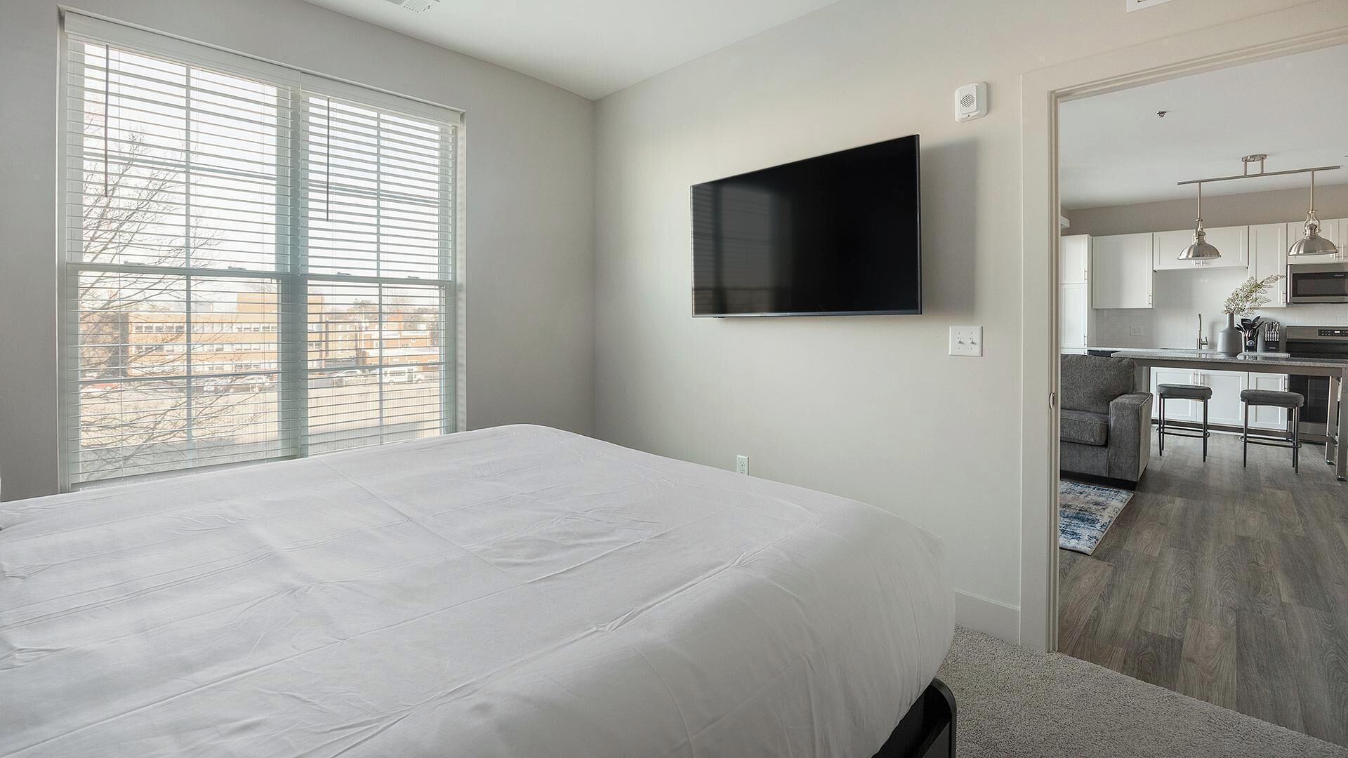 Une chambre moderne avec un grand lit, un téléviseur fixé au mur et une vue sur la cuisine et le salon, grâce à une porte ouverte.