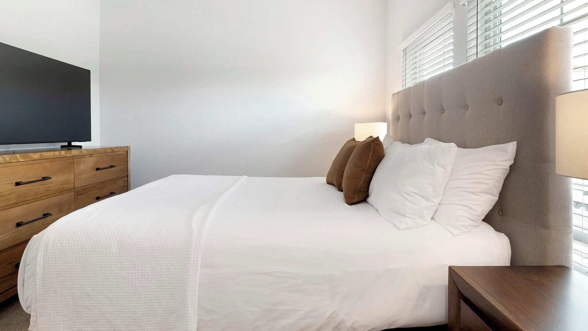 Ein ordentlich bezogenes Bett mit weißen Leinentüchern, eine hölzerne Kommode mit Schubladen, ein Fernseher darauf und ein Fenster mit Jalousien.