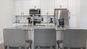 A modern kitchen with white cabinets, stainless steel appliances, granite countertops, and three grey chairs. - Landing | Stylish 1BD, Pool, Gym (Palm Bay)