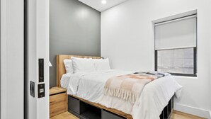Quarto com cama, mesa de cabeceira, janela com persianas e gavetas embutidas sob a cama.
