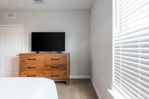 A bedroom with a bed, a wooden dresser, a television on top, and a window with blinds. - Landing | Stylish 2BD, Pool, Gym (Palm Bay)
