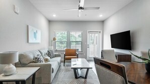 Un salon moderne avec un canapé, une table basse et des fauteuils, un téléviseur à écran plat fixé au mur et un ventilateur de plafond.